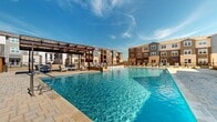 This two-floor residential community centers around a standout resort-style outdoor amenity space designed for leisure and relaxation. The centerpiece is a generously sized swimming pool with crystal-clear blue water, framed by a wide stone-paved sundeck lined with lounge chairs — ideal for sunbathing and outdoor recreation. A cascading water feature adds a dynamic architectural element along one side of the pool, enhancing the ambiance with both visual appeal and the soothing sound of flowing water. A pergola structure on one end of the deck provides shaded seating, offering a comfortable gathering space for residents. Flanking the pool area is a beautifully designed clubhouse building, constructed with light brick and featuring floor-to-ceiling windows that allow abundant natural light to fill the interior while offering views of the pool and surrounding landscape. The exterior of the clubhouse opens onto a spacious paver patio, extending the usable outdoor area. The overall architectural style of the community blends contemporary and traditional design elements, with multi-story residential buildings in neutral earth tones providing an attractive backdrop to the outdoor amenity area. The open sky above and broad outdoor sightlines create a bright, airy atmosphere throughout the entire space.