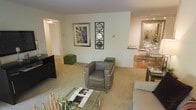 This single-floor residence offers an open and functional layout that seamlessly connects the living and dining areas. The living room is anchored by plush, wall-to-wall carpeting in a warm neutral tone, creating a comfortable foundation for the contemporary furnishings. A sleek leather armchair with chrome accents serves as a focal point, complemented by green upholstered ottomans and a glass-top coffee table that add visual interest without overwhelming the space. Decorative wall art, including a framed cityscape photograph and a geometric mirror panel, lend a modern, curated aesthetic to the cream-colored walls. A dark entertainment console positioned along one wall provides practical media storage. Adjacent to the living area, the dining space transitions to a hard surface floor and features a glass dining table with metallic chairs, adding an elegant touch. Track lighting overhead illuminates the dining area effectively. A hallway visible from the living room leads to additional rooms, suggesting a well-organized floor plan. Overall, the space presents a clean, contemporary ambiance with a cohesive neutral palette accented by green and metallic tones.