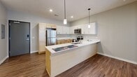 This single-story space is designed with an open-concept layout that seamlessly connects the kitchen and living area, creating a cohesive and functional floor plan. The kitchen is a central highlight, featuring a large island with a white countertop that doubles as a breakfast bar, dual pendant lights overhead, and a stainless steel appliance suite including a refrigerator, range, microwave, and dishwasher. Cabinetry combines light natural wood tones on the lower sections with crisp white upper cabinets, offering ample storage throughout.

Wood-look flooring flows consistently across the entire unit, lending warmth and visual continuity to each room. The living area is open and well-lit, benefiting from a large sliding door dressed with vertical blinds that allows natural light to fill the space. Recessed lighting and a textured ceiling complement the neutral gray walls found throughout.

The bedroom is a comfortable, well-proportioned room with a ceiling fan, a window with blinds, and generous natural light. Two bathrooms maintain the same clean aesthetic, each featuring a light wood vanity with a white countertop and wood-look flooring. The overall ambiance is clean, contemporary, and move-in ready.