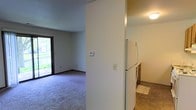 This single-floor residential space offers a straightforward and functional layout designed for comfortable everyday living. The living area is finished with wall-to-wall carpeting, creating a warm and inviting atmosphere, and is bathed in natural light courtesy of a large sliding glass door that opens to an outdoor view of lush greenery. The neutral-toned walls throughout lend a clean, versatile aesthetic that easily accommodates a variety of interior styles.

A partial wall divides the living area from the kitchen, providing a sense of separation while maintaining an open feel between the two spaces. The kitchen features tile flooring, offering a practical and easy-to-maintain surface. It is equipped with a full-size refrigerator and cabinetry that provides ample storage for everyday needs. Overhead lighting in the kitchen ensures the space is well-lit for meal preparation.

A thermostat is conveniently mounted on the dividing wall, allowing for easy climate control throughout the unit. The overall design is modest and efficient, prioritizing functionality with a clean, neutral palette. The combination of carpeted living spaces and tiled kitchen areas reflects a thoughtful approach to practicality, making this a well-suited space for a range of occupants.