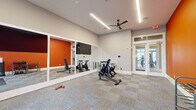 This dedicated fitness room offers a thoughtfully designed workout environment within a compact yet efficient footprint. The space features soaring ceilings that contribute to an open, airy atmosphere, complemented by multiple windows that flood the room with natural light and offer views of the surrounding greenery. A ceiling fan overhead ensures comfortable airflow during workouts.

The interior design is defined by bold, energizing orange and red accent walls set against neutral gray tones, creating a motivating aesthetic. A large wall-mounted mirror spans one side of the room, visually expanding the space while allowing users to monitor their form. Durable, multi-tone carpet tiles cover the floor throughout, providing both cushioning and texture suited to a variety of exercise activities.

Equipment includes stationary spin bikes, an adjustable workout bench, a cable resistance machine, a dumbbell rack with free weights, kettlebells, medicine balls, and yoga mats — catering to cardio, strength, and flexibility training alike. A wall-mounted flat-screen TV adds an entertainment element to workouts. Glass-paned double doors provide access to an adjacent fitness area, suggesting this room is part of a broader amenity facility. Linear LED lighting fixtures mounted to the ceiling ensure bright, even illumination throughout the space.