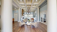 Located at Pointe of Woods in Sayreville, NJ, this sophisticated community clubhouse is a masterclass in classical design and refined detailing. The grand entrance foyer welcomes visitors with soaring ceilings, stately columns, a sweeping arched window, and rich hardwood flooring that flows throughout the space. A dramatic chandelier anchors the entryway, setting an elegant tone from the moment you arrive.

The expansive living room serves as the centerpiece of the space, highlighted by a floor-to-ceiling natural stone fireplace flanked by built-in shelving and cabinetry. Coffered ceilings add architectural depth, while an abundance of windows and French doors bathe the room in natural light and provide direct views of the outdoor grounds. Tall decorative columns define the space while maintaining an open, airy feel.

Adjacent to the living area, a well-appointed lounge features a built-in credenza, arched mirrors, and additional French doors that open to the exterior, further enhancing the connection to the outdoors.

The dining room is distinguished by soft sage-toned wainscoting, tall ceilings with crown molding, and generous natural light from multiple windows. Round dining tables provide a casual yet polished setting, and a doorway connects seamlessly back to the main living and entry areas, ensuring smooth flow throughout this thoughtfully designed communal space.