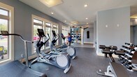 This fitness center in Clover, SC is a thoughtfully designed, single-floor facility built to support a wide range of workout routines. The space is finished with soft blue-gray walls, white trim, and durable rubber-backed flooring, creating a clean and motivating environment throughout.

The cardio zone is well-appointed with multiple treadmills, elliptical trainers, and an air resistance bike, all positioned along walls lined with large windows that flood the room with natural light and offer views of the surrounding outdoor greenery. A wall-mounted flat-screen television adds entertainment options for endurance training sessions.

The strength training area includes a multi-functional cable machine, a dedicated rowing machine, a weight bench, and a dumbbell rack, providing comprehensive options for resistance-based workouts. A functional training station features a wall-mounted pull-up and suspension training rig alongside a heavy punching bag, catering to athletic and cross-training enthusiasts.

Recessed lighting throughout supplements the natural light, ensuring the space remains bright and well-lit at all times. The open layout, supported by structural columns, allows for easy movement between equipment zones. Large glass panel windows and doors connect the fitness area visually to adjacent spaces, contributing to an airy and expansive feel. Overall, this facility delivers a complete and well-organized fitness experience.