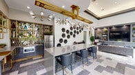 Located at 3820 W Northwest Highway in Dallas, Texas, this single-story clubhouse at Elan Apartments presents a sophisticated, contemporary design that blends bold aesthetics with functional community amenities. The space is organized into distinct yet open zones that flow seamlessly from one to the next.

The entrance lobby immediately establishes the design tone, featuring polished marble-look flooring, soaring floor-to-ceiling glass walls, and a branded welcome mat flanked by accent chairs and lush tropical greenery. The transparency of the entryway fills the space with abundant natural light while offering views to the surrounding exterior.

Moving deeper into the clubhouse, a striking kitchen and bar area serves as a central focal point. A large granite-topped island with quilted leather barstools is anchored beneath gold-framed pendant chandeliers with globe bulbs. Mirrored hexagonal tile backsplash, stainless steel appliances, and gold-accented cabinetry complete the refined look.

Adjoining the kitchen, a spacious lounge area features eclectic décor, including sculptural wall art, a vintage-style scooter display piece, tufted velvet seating, and a live-edge wood conference table. Floor-to-ceiling windows frame direct views of the outdoor pool and courtyard, further enhancing the open, airy ambiance throughout this well-appointed community space.