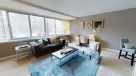 This single-floor space is designed to balance comfort and functionality within a well-organized open layout. The living area serves as the centerpiece, anchored by a charcoal sofa adorned with teal and mustard accent pillows, positioned atop a vibrant blue area rug that adds a bold pop of color to the neutral palette. A sleek, dark rectangular coffee table sits at the heart of the seating arrangement, while a patterned accent chair and metallic side accents contribute texture and visual interest.

Large windows span one wall, flooding the space with natural light and framing views of mature trees outside, creating a connection to the outdoors. Warm wood-toned flooring and soft greige walls provide a calm, neutral backdrop throughout.

A dedicated workspace is seamlessly integrated into the room, featuring a compact light-wood desk, an ergonomic chair, and a task lamp — ideal for working from home. Wall-mounted framed artwork above the desk adds a curated, polished touch. The overall ambiance is modern and inviting, with a thoughtful mix of functional furnishings and contemporary décor that maximizes the utility of every corner.