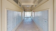 The mail area is a single-story, open-ended corridor designed to serve as a central hub for resident mail collection. The passageway is lined on both sides with rows of aluminum individual mailboxes, offering organized and secure mail access for a large number of residents. The flooring features a brick-patterned tile in warm neutral tones, adding a structured yet inviting aesthetic to the functional space.

The corridor benefits from natural light streaming in through glass-panel doors and open entryways at both ends, creating a bright and airy atmosphere. The ceiling is finished with light-toned wood paneling, complementing the warm beige walls and giving the space a clean, cohesive look. Upper transom windows further enhance the flow of natural light throughout the corridor.

At one end, the space opens toward a landscaped outdoor courtyard area featuring greenery, a waste receptacle, and a bench for resident convenience. The opposite end opens to a parking or exterior access area. A bulletin board display case is mounted near the mailboxes, providing space for community notices. Overall, the mail area is thoughtfully designed to balance practicality with a welcoming environment.