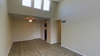 This single-story space is defined by its dramatically high ceilings, which lend an airy, expansive quality to the interior. A row of clerestory windows positioned near the top of the main living area allows abundant natural light to pour in, illuminating the neutral beige walls and contributing to an open, welcoming ambiance. The flooring throughout is wood-style plank, offering a warm, cohesive look that complements the light-toned walls.

The main living area is generously proportioned, with a ceiling fan fixture installed in a lower-ceiling section near the entryway, providing both functionality and subtle character. The front entry features a solid white door and transitions smoothly into the living space. A recessed niche in one wall adds an architectural detail that breaks up the clean lines of the room.

Adjacent to the living area, a partially visible kitchen space features dark cabinetry, offering a contrast to the otherwise light interior palette. The open passage between spaces supports a connected flow throughout the unit. Overall, the space presents a clean, practical design with notable vertical volume and ample natural light as its defining characteristics.