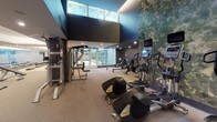 Located at 1201 Half St SE in Washington, DC, this contemporary fitness center is thoughtfully designed to accommodate a wide range of workout routines. The facility is divided into distinct functional zones, each outfitted with professional-grade equipment. The cardio area features a row of treadmills and elliptical machines positioned against a striking botanical-print accent wall, creating an immersive and visually engaging environment. Nearby, a dedicated strength training zone houses multiple power cages, free weights, benches, and cable machines, all set against floor-to-ceiling mirrors that amplify the sense of space. A collection of stability balls, medicine balls, and functional fitness accessories rounds out the strength offerings. A separate group fitness studio features light hardwood flooring, full-length mirror walls, and large windows that invite natural light — Reebok aerobic step platforms are arranged throughout, making the room well-suited for structured classes or solo training. The facility benefits from soaring double-height ceilings in key areas, floor-to-ceiling glazing with outdoor views, and a sophisticated dark-toned color palette accented by recessed and cove lighting. Frosted glass partitions provide access to additional support spaces, completing a well-appointed, modern fitness experience.