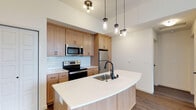 This single-floor residence showcases a thoughtfully designed open-concept layout with a strong emphasis on modern finishes and functional living. The kitchen serves as the heart of the space, featuring warm natural wood cabinetry that provides ample storage, complemented by a classic white subway tile backsplash that adds a clean, timeless aesthetic. Stainless steel appliances, including a French door refrigerator, electric range, and over-the-range microwave, outfit the kitchen with everything needed for everyday cooking. A standout feature is the generously sized curved kitchen island, finished with a crisp white quartz countertop and an undermount stainless steel sink with a matte black faucet, creating an elegant focal point ideal for both meal preparation and casual dining. Durable wood-look flooring flows throughout, grounding the space with a warm, cohesive tone. Lighting is a key design element, with a combination of a contemporary ceiling fixture and a trio of industrial-style mason jar pendant lights suspended above the island, adding character and charm. The open layout and light neutral wall tones contribute to an airy, welcoming ambiance throughout the space.