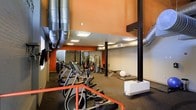 This fitness center offers a spacious, single-floor layout designed to accommodate a full range of workout activities. The space is defined by its industrial aesthetic, highlighted by exposed ductwork running along the upper walls and structural support columns painted in a contrasting dark tone. Bold orange accent walls add energy to the environment, complementing the high ceilings that contribute to an open, airy atmosphere.

The cardio zone is prominently featured near the entrance, with a row of elliptical machines and other cardio equipment arranged for easy access. Further into the space, a dedicated strength training area includes a multi-station cable and weight machine setup, free weights, and exercise balls, offering a comprehensive range of resistance training options. Rubber flooring throughout provides safety and durability across all workout zones.

Large wall-mounted mirrors in the strength area enhance the sense of depth and allow users to monitor their form during exercises. Recessed and flush-panel ceiling lighting ensures the entire space is well-lit. The layout is thoughtfully organized to separate cardio and strength training areas while maintaining an open flow, making this fitness center a functional and motivating environment for users of all fitness levels.