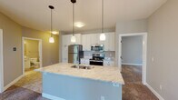 This single-floor residence is designed with an open-concept layout that seamlessly connects the living area to the kitchen. The kitchen is well-appointed with white shaker-style cabinetry offering ample storage, stainless steel appliances including a refrigerator, range, and microwave, and a light granite-topped island with a double sink. Pendant lights hang above the island, complementing the overall warm, neutral color palette found throughout the unit.

The living area is spacious and carpeted, featuring a curved bay window arrangement that invites abundant natural light and outdoor views, alongside a sliding glass door that opens to a balcony. A ceiling fan provides additional comfort to the space.

The bedrooms are carpeted and finished in warm neutral tones, each equipped with ceiling fans. One bedroom features a walk-in closet with built-in shelving, providing practical storage solutions.

Two bathrooms serve the unit, both finished consistently with white cabinetry, granite countertops, and tile flooring. The primary bathroom includes a walk-in shower with glass doors, while the secondary bathroom offers a shower-tub combination. Both feature large mirrors and adequate vanity space.