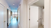 This thoughtfully designed one-bedroom, one-bathroom unit offers a well-organized single-floor layout that maximizes every square foot. The interior is defined by a clean, contemporary aesthetic featuring white walls, recessed lighting, and dark hardwood-style flooring throughout.

The open-concept living and kitchen area serves as the heart of the unit. The living space accommodates a comfortable seating arrangement anchored by a soft area rug, a sleek media console, and an accent chair in warm amber tones that adds a pop of color. Floor-to-ceiling windows flood the space with natural light and provide views of the surrounding urban environment. The kitchen is neatly integrated with light cabinetry, built-in appliances, and a clean countertop workspace.

A hallway connects the main living area to the private bedroom, which is generously proportioned for the unit's overall scale. The bedroom features neutral walls, recessed lighting, and a large window that continues the theme of natural light. Abstract wall art adds a warm, curated touch.

The bathroom is finished with large-format beige tiles on both the floor and walls, a floating wood-toned vanity with a white countertop, and a full tub-shower combination enclosed by a curtain. Brushed nickel hardware and grab bars provide both style and practicality throughout.