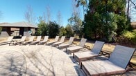 Located in Columbus, Georgia, this outdoor amenity space showcases a beautifully designed resort-style pool and expansive patio area. The centerpiece is a large, freeform swimming pool with a gradual entry, its clear waters reflecting the surrounding natural scenery. The pool's curved edges and stone coping contribute to an elegant, inviting aesthetic.

Surrounding the pool, a generously sized stone-paved patio provides ample room for relaxation and socializing. Numerous sling-style chaise lounges with wood frames are arranged throughout the deck, offering comfortable spots to lounge in the Georgia sunshine. Teal patio umbrellas provide shaded seating areas, adding a pop of color against the neutral stone and dark exterior of the main building.

The backdrop features a striking multi-story structure with a stone and dark-siding exterior, accented by large windows and a covered balcony, lending an upscale architectural presence to the space. Mature evergreen and deciduous trees frame the perimeter, creating a natural privacy buffer and a serene, park-like atmosphere. The combination of refined hardscaping, thoughtful furnishings, and abundant greenery makes this outdoor space a standout feature of the property.