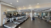 The fitness center at 101 Washington Blvd in Stamford, Connecticut is a thoughtfully designed, single-floor facility offering a comprehensive workout environment. The space is organized into distinct functional zones, each catering to different aspects of physical training. The main cardio area features a row of modern treadmills lined along large wall-to-wall mirrors that visually expand the space and allow users to monitor their form during exercise. Recessed ceiling lighting and a neutral warm color palette create a clean, focused atmosphere throughout. A dedicated cardio alcove benefits from floor-to-ceiling windows on multiple walls, flooding the area with abundant natural light and views of the surrounding streetscape. This section houses elliptical trainers, upright and recumbent stationary bikes, and a stair climber. A separate strength training zone is equipped with a multi-station cable machine, rowing equipment, and free weights, supported by instructional wall charts and a stability ball for functional fitness. Durable rubber flooring covers the entire space, providing safety and noise reduction. Colorful abstract artwork accents several walls, adding visual energy to the environment. The facility is well-ventilated with visible HVAC ductwork and climate control systems, ensuring comfort during intensive workouts.