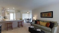 Located at 5885 Tuttle's Grove Blvd in Dublin, Ohio, this single-floor residence offers a thoughtfully designed open-concept layout that seamlessly connects the main living areas. The living room is a welcoming space, highlighted by plush carpeted flooring, neutral-toned walls, and a ceiling fan that adds both function and visual interest overhead. A tufted ottoman coffee table and a comfortable sofa anchor the room, while a framed wall accent adds a decorative focal point.

The living area flows naturally into a dedicated dining space, which accommodates a round dining table with traditional Windsor-style chairs. A large window dressed with vertical blinds allows generous natural light to fill the room, creating a bright and airy atmosphere throughout.

The kitchen is connected to the living and dining areas via a convenient pass-through counter with bar stool seating, making it ideal for casual dining or entertaining. The kitchen features white cabinetry and offers a functional workspace. Visible HVAC venting integrated into the ceiling ensures year-round climate comfort. The overall design of this single-floor home emphasizes an open, cohesive flow between spaces, making it a practical and comfortable residence in the Dublin, Ohio community.