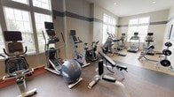 This fitness center offers a thorough and well-organized workout environment spread across a single floor. The space is thoughtfully divided into dedicated zones for cardio, free weights, and strength training, accommodating a wide range of exercise routines. Large windows line the walls throughout, flooding the interior with abundant natural light and providing views of the surrounding outdoors, creating an energizing atmosphere for workouts. The walls feature a distinctive grey horizontal stripe accent against a neutral palette, lending the space a clean, modern aesthetic. Recessed lighting supplements the natural light, ensuring the facility is well-lit at all times. The cardio zone is equipped with multiple treadmills, elliptical trainers, and stationary and spin bikes, many featuring individual display monitors. The free weight area includes a fully stocked dumbbell rack alongside adjustable benches and barbell stations suited for a variety of lifts. The strength training section houses cable machines and multi-functional weight systems, supported by stability balls and medicine balls for functional fitness. Durable rubber flooring covers the high-traffic workout areas, transitioning to a neutral carpet near the perimeter. French doors provide easy access to an adjacent entryway. Framed artwork on the walls adds a polished, welcoming touch to the overall environment.