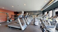 Located at 101 Summit St in Duluth, MN, this fitness center is a comprehensively outfitted single-room exercise facility designed to support a full range of workout routines. The space is organized into distinct cardio and strength training zones, making efficient use of its footprint across a single floor.

The cardio area is lined with a row of Precor treadmills and elliptical machines, each equipped with individual display screens. Large mirrors run along one wall, amplifying the sense of space and allowing users to monitor their form. Windows along the cardio side bring in natural light and offer exterior views, enhancing the workout environment. A wall-mounted fan provides additional air circulation throughout the space.

The strength training section features a cable cross machine, a squat rack with weight plates, a full dumbbell rack ranging across multiple weights, and a flat bench, providing ample options for resistance training. A wall-mounted television is positioned above the strength area for entertainment during workouts.

The interior design employs a bold two-tone color scheme, with deep blue-gray accent walls on one side and warm coral-orange walls on the other, creating an energetic and motivating atmosphere. The durable speckled rubber flooring is practical and well-suited for high-traffic use. Recessed lighting and exposed ductwork along the ceiling contribute a modern, industrial aesthetic. Framed athletic photography decorates the walls, adding to the facility's focused fitness ambiance.