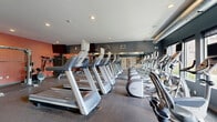 Located at 101 Summit St in Duluth, MN, this fitness center is a comprehensively outfitted single-room exercise facility designed to support a full range of workout routines. The space is organized into distinct cardio and strength training zones, making efficient use of its footprint across a single floor.

The cardio area is lined with a row of Precor treadmills and elliptical machines, each equipped with individual display screens. Large mirrors run along one wall, amplifying the sense of space and allowing users to monitor their form. Windows along the cardio side bring in natural light and offer exterior views, enhancing the workout environment. A wall-mounted fan provides additional air circulation throughout the space.

The strength training section features a cable cross machine, a squat rack with weight plates, a full dumbbell rack ranging across multiple weights, and a flat bench, providing ample options for resistance training. A wall-mounted television is positioned above the strength area for entertainment during workouts.

The interior design employs a bold two-tone color scheme, with deep blue-gray accent walls on one side and warm coral-orange walls on the other, creating an energetic and motivating atmosphere. The durable speckled rubber flooring is practical and well-suited for high-traffic use. Recessed lighting and exposed ductwork along the ceiling contribute a modern, industrial aesthetic. Framed athletic photography decorates the walls, adding to the facility's focused fitness ambiance.