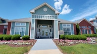 This single-story building presents a warm and inviting first impression with its coastal-inspired architectural design. The exterior showcases sage green horizontal lap siding complemented by brick accents, a pitched gable roofline, and a distinctive circular vent detail centered above the entrance. A covered front porch welcomes visitors with classic white rocking chairs flanking a set of elegant glass-paneled double doors, creating an approachable and relaxed atmosphere.

The building's entrance is framed by a well-maintained landscape featuring colorful flower beds with pink and white blooms, neatly trimmed boxwood shrubs, and lush green grass. A concrete walkway leads directly to the front entrance, providing a clean and accessible approach. Transom windows above the entry doors and additional windows along the facade allow natural light to fill the interior while maintaining architectural charm.

The overall ambiance of this space is one of community and comfort, blending traditional Southern design elements with a light, airy color palette. The thoughtfully maintained grounds and classic architectural details convey a sense of quality and attention to detail, making this an attractive and functional gathering or welcoming space for its community.