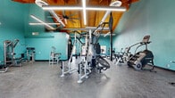 Located at 370 E Harmon Ave in Las Vegas, NV, this fitness center offers a generously sized, single-floor workout space designed to accommodate a comprehensive range of exercise needs. The room's most distinctive architectural feature is its dramatic vaulted ceiling, where warm-toned exposed wood beams and tongue-and-groove paneling create an inviting, open atmosphere. Large industrial ceiling fans and linear LED lighting run the length of the ceiling, ensuring the space remains bright and well-ventilated throughout. Exposed ductwork adds an industrial touch that complements the overall aesthetic. The walls are finished in a bold teal, lending a vibrant and energizing ambiance to the space. Durable speckled concrete flooring spans the entire area, providing a practical and easy-to-maintain surface suited for high-traffic use. The fitness floor is outfitted with a diverse selection of equipment, including multi-station cable machines, weight benches, selectorized strength machines, a heavy punching bag, treadmills, ellipticals, stationary bikes, and stair climbers. Large windows along the perimeter allow natural light to filter in and offer views of the surrounding outdoor landscaping, including palm trees. A glass double-door entrance at the front welcomes visitors and further enhances the sense of openness throughout this well-appointed facility.