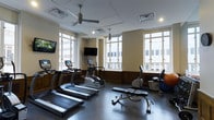 This fitness center offers a thoughtfully designed workout space that balances functionality with a welcoming ambiance. The room is anchored by large, multi-pane windows that line the walls, allowing abundant natural light to fill the space while offering views of the surrounding urban environment. The ceiling features ceiling fans and recessed lighting, ensuring proper air circulation and consistent illumination throughout the facility. The flooring is a durable, dark rubber material well-suited for high-traffic exercise use. The cardio area is well-appointed with a selection of treadmills, an elliptical trainer, a stationary upright bike, and a recumbent bike, catering to a range of workout preferences. A weight bench and exercise ball round out the equipment offerings, providing options for strength and flexibility training. Two wall-mounted flat-screen televisions are positioned for easy viewing from multiple points in the room, enhancing the exercise experience. The warm-toned walls and classic wainscoting detailing give the space a refined character that sets it apart from a typical gym environment, creating an inviting atmosphere that encourages regular use.
