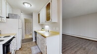 This single-floor unit is thoughtfully laid out across several distinct areas, each with its own character and function. The galley-style kitchen is efficiently organized, featuring white cabinetry with decorative wire-mesh upper doors, granite-look countertops, a stainless steel sink, and white appliances including an electric range and refrigerator. Durable wood-look vinyl plank flooring runs throughout the kitchen and connects seamlessly to the adjoining living spaces.

The open living area is bright and spacious, benefiting from a large sliding glass door that floods the room with natural light and provides access to an outdoor space. The neutral-toned walls, textured ceiling, and gray-toned vinyl plank flooring give the room a clean, versatile aesthetic. A baseboard heating unit runs along one wall. A secondary open area near the entryway features a built-in closet with louvered doors, offering convenient storage.

The bedroom is a calm, private retreat with soft carpeting, warm neutral walls, a textured ceiling, a window with blinds, and a baseboard heater. The bathroom includes a vanity with a decorative cabinet, a stone-patterned countertop, an undermount sink, and classic white tile wall surround, completing the unit with a timeless, functional finish.