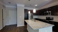 Located at 3112 Maybank Hwy on Johns Island, South Carolina, this single-story residence showcases a thoughtfully designed interior with contemporary finishes throughout. The kitchen serves as a central highlight, featuring an expansive granite island with an integrated sink and pendant lighting overhead, creating both a functional workspace and an inviting gathering area. Dark espresso cabinetry extends to the ceiling, providing generous storage while lending a bold, sophisticated contrast to the light granite countertops and neutral gray walls. A classic white subway tile backsplash adds a clean, timeless detail behind the counters. Stainless steel appliances, including a full-size refrigerator and built-in microwave, complete the kitchen's polished aesthetic. Crown molding and decorative column details lend an architectural refinement to the space, while dark hardwood-style flooring runs throughout, grounding the interior with warmth and continuity. The overall ambiance is modern and well-appointed, balancing dark and light tones to create a space that feels both stylish and functional.
