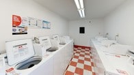 This shared laundry room is a well-organized, single-floor facility designed for practical, everyday use. The space is immediately recognizable by its bold red-and-white checkered vinyl flooring, which runs throughout and gives the room a clean, classic aesthetic. Bright fluorescent lighting installed along the ceiling ensures the entire area is well-lit at all times.

Rows of top-loading washing machines line both sides of the room, providing ample capacity for multiple users. A set of dryers is also present, positioned alongside the washers for a convenient, streamlined workflow. Instructional and safety notices are posted on the walls near the machines for easy reference.

A built-in folding counter extends along one wall, offering a dedicated surface for sorting and folding laundry. A red utility cabinet provides additional storage within the space. A wall-mounted fire extinguisher box and a ventilation panel are also present, reflecting attention to safety and air circulation.

Multiple windows allow natural light to enter and offer views of the surrounding outdoor greenery, creating a more pleasant environment. A glass door provides direct exterior access, further enhancing ventilation and ease of entry. The white walls and bright finishes contribute to an overall clean and functional ambiance.