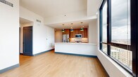 This well-appointed one-bedroom, one-bathroom residence offers a thoughtfully designed layout across a single floor, combining modern finishes with functional living spaces. The open-concept kitchen and living area serves as the heart of the home, featuring light hardwood flooring, bold navy baseboards, and large floor-to-ceiling windows that frame impressive city skyline views and flood the space with natural light. The kitchen is equipped with stainless steel appliances, including a gas range, built-in microwave, refrigerator, and dishwasher. Warm wood cabinetry in two complementary tones pairs with crisp white countertops and a subway tile backsplash, while statement dome pendant lights add a contemporary touch above the peninsula. The bedroom is carpeted for comfort and benefits from expansive windows with striking urban views, and features a stylish sliding barn door leading to a dedicated closet. The full bathroom is finished with stone-look tile flooring, a sleek vanity with a white countertop, a framed mirror with warm-toned hardware, and a glass-enclosed shower. A clean, well-lit hallway connects the rooms, finished with the same light wood flooring and recessed lighting seen throughout the common areas, completing the cohesive, modern aesthetic of this urban residence.