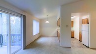 This single-floor space features an open-concept layout that seamlessly connects the living, dining, and kitchen areas. The dining area is highlighted by a classic pendant light fixture suspended from the ceiling, adding a focal point to the space. Neutral-toned carpet covers the living and dining areas, contributing to a warm and understated aesthetic throughout.

Natural light filters in through a large window dressed with vertical blinds, brightening the main living area. A sliding glass door on the adjacent wall offers additional light and potential access to an outdoor area. The walls are finished in a clean, neutral tone that enhances the sense of openness.

The kitchen is partially separated by a half-wall partition, creating a defined yet connected cooking space. It features warm wood-toned cabinetry providing ample storage, along with a countertop that extends along the perimeter. The kitchen floor transitions to a hard surface material, offering a practical contrast to the carpeted living spaces. Overall, the layout is functional and straightforward, suited for everyday living.