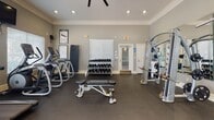 Located at 1101 Luther St W in College Station, Texas, this fitness center offers a well-equipped, single-floor workout facility designed to support a full range of exercise routines. The space is finished with neutral gray walls, crown molding, and dark rubber flooring, creating a clean and functional environment. Recessed lighting and a ceiling fan help maintain comfort throughout the room.

The cardio area features treadmills, stationary cycling bikes, and a rowing machine, all positioned along a windowed wall fitted with white horizontal blinds that allow natural light to filter in. Wall-mounted flat-screen televisions are installed above the cardio equipment, providing entertainment during workouts.

The strength training area is equipped with multi-station cable machines, a cable crossover unit, and a selection of selectorized weight machines. A full rack of dumbbells lines one wall, accompanied by adjustable weight benches to support free weight exercises. Large wall-to-wall mirrors span the strength training area, visually expanding the space and allowing users to monitor their form.

Additional amenities include a dedicated exercise mat for stretching or floor exercises and informational workout charts posted on the wall. The facility is bright, organized, and thoughtfully arranged to accommodate multiple users simultaneously, offering a comprehensive fitness experience.
