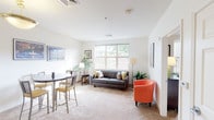 This single-floor residence offers a thoughtfully designed layout that balances comfort and functionality. The open living area is bathed in natural light from a large multi-pane window, creating a warm and inviting atmosphere. Neutral-toned carpet and crisp white walls provide a clean backdrop, while the space comfortably accommodates a tufted sofa, accent chair, and side seating area. A dedicated dining nook sits adjacent to the living area, featuring a round table and chairs beneath framed cityscape artwork, with ample natural light adding to the airy feel. The galley-style kitchen is efficiently organized with warm wood cabinetry, tile flooring, and full appliances including a refrigerator, range, and dishwasher, offering generous counter and storage space. The bedroom is a serene retreat with carpeted floors, a large window with blinds, and a walk-in closet visible through the doorway, providing substantial storage. The bathroom is designed with accessibility in mind, featuring grab bars, a shower-tub combination with a curtain, tile flooring, a vanity with ample counter space, and a well-lit mirror. Throughout the residence, neutral tones and practical finishes create a cohesive and comfortable living environment.
