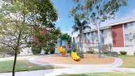 This outdoor playground area is situated within a landscaped courtyard, offering a dedicated recreational space designed for children. The play area centers around a multi-component structure finished in bold yellow and blue, featuring a prominent spiral slide that curves dramatically from an elevated platform to the ground below. A secondary straight slide and climbing elements provide additional play options, catering to a range of ages and activity levels.

The playground surface is covered with wood chip mulch, providing a safety-conscious ground covering beneath and around the equipment. The space is bordered by a smooth concrete pathway that curves around the perimeter, creating clear separation between the play zone and surrounding landscaped areas. Mature trees, including a large eucalyptus and a palm tree, offer natural shade and visual character to the courtyard.

The surrounding buildings, finished in neutral tones with structured facades, frame the space on multiple sides, giving the courtyard an enclosed and sheltered feel. Trimmed shrubs and green plantings line the building edges, contributing to a well-maintained appearance. The overall setting benefits from open sky above, ensuring ample natural light throughout the day.
