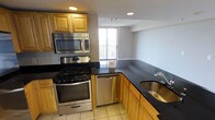 This single-floor space centers around a thoughtfully designed kitchen that balances functionality with style. The kitchen features warm honey-toned wood cabinetry that provides ample storage, complemented by striking dark granite countertops that run along an L-shaped layout, offering generous prep and workspace. Stainless steel appliances include a gas range, over-the-range microwave, dishwasher, and a double-basin sink, creating a cohesive and modern aesthetic throughout. Light-toned tile flooring adds brightness to the space while providing a durable, easy-to-maintain surface. Recessed lighting in the ceiling ensures even illumination throughout the kitchen. A standout feature is the large sliding glass door at the far end of the space, which floods the area with natural light and provides direct access to an outdoor balcony, enhancing the sense of openness. The layout flows naturally from the kitchen into an adjacent area, making the overall space feel connected and practical. The neutral gray wall tones throughout complement the warm wood and dark stone finishes, creating a balanced and inviting atmosphere that is both functional and visually appealing.