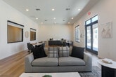 Located at 18730 Miner Ln in Morgan Hill, California, this single-floor community space offers a thoughtfully designed open-plan living area that blends modern aesthetics with functional comfort. The interior is finished with light hardwood flooring that runs throughout, contributing to a warm yet contemporary atmosphere.

The main living area is generously proportioned, anchored by a large, neutral-toned sofa ensemble positioned on a patterned area rug. A striking feature wall with vertical shiplap-style paneling serves as a focal point, complete with a wall-mounted flat-screen television. Complementary seating, including a plush accent chair and a sleek coffee table, complete the lounge arrangement.

Toward the back of the space, a round dining table with upholstered chairs provides a dedicated seating zone, ideal for small group gatherings. Adjacent to this area, a wet bar or kitchenette with dark cabinetry and a countertop adds practical functionality. The walls are finished in crisp white, enhancing the sense of openness and brightness.

Natural light flows in through multiple large black-framed windows and a set of French doors, offering views to the exterior and creating an airy, inviting ambiance. Recessed ceiling lighting complements the natural light, ensuring the space is well-lit throughout the day.
