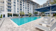 Located at 3955 Design Center Dr in Palm Beach Gardens, Florida, this resort-style outdoor amenity space showcases a generously sized swimming pool as its centerpiece, framed by a sleek, modern multi-story residential building. The pool area is designed with a contemporary aesthetic, featuring clean geometric lines, light-toned stone pavers, and crystal-clear turquoise water that evokes a luxury resort atmosphere. Surrounding the pool, an expansive sun deck is furnished with rows of white lounge chairs and shaded by large navy-blue market umbrellas, offering ample space for relaxation. Lush tropical landscaping, including towering queen palms and ornamental plantings, enhances the Florida ambiance throughout the space. Dedicated lounge seating areas with cushioned sofas and accent chairs are positioned poolside, creating comfortable social gathering spots. A pergola-covered outdoor living area with a television provides a shaded retreat, while multiple open lounging zones offer flexibility for sunbathing. The pool features varying depths, with clearly marked shallow and deep ends, and is bordered by integrated step entries. The surrounding multi-story building features floor-to-ceiling windows and private balconies that overlook the pool deck, completing this well-appointed outdoor amenity space.