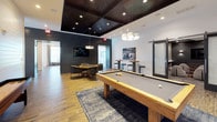 Located at 1605 Barclay Pointe in Wilmington, North Carolina, this single-floor amenity space is thoughtfully designed to offer a blend of entertainment and relaxation. The game room serves as a central attraction, showcasing a solid wood pool table set atop a layered area rug over warm hardwood-style flooring. A shuffleboard table lines the shiplap-clad white wall, while high-top bar tables with stools provide casual seating nearby. The room's dark, coffered wood ceiling with recessed and pendant lighting creates a striking contrast against the bright white shiplap walls and bold navy accent wall, which is decorated with sculptural metallic wall art. A wall-mounted flat-screen TV and cozy lounge seating round out the space. A sliding barn door leads to a connected media lounge, which features a sophisticated dark gray palette, textured carpet, and a large wall-mounted television flanked by geometric sconce lighting. Plush curved lounge chairs paired with round wood side tables and powder-blue ottomans create a comfortable theater-like arrangement. Nautical-themed artwork throughout ties the design together, nodding to Wilmington's coastal character. The overall ambiance is modern yet inviting, with a refined coastal aesthetic.
