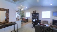 This single-floor space showcases an open-concept layout that effortlessly connects the living and dining areas into one cohesive, flowing environment. The vaulted ceiling is a defining architectural feature, adding a sense of height and airiness throughout the main living area. Recessed lighting is installed throughout the ceiling, providing even, ambient illumination that complements the abundant natural light streaming in through multiple windows positioned along the walls.

The living area is anchored by a comfortable seating arrangement centered around dark leather chairs and a sofa, with a tall, dark cabinet serving as a functional and decorative focal point along one wall. A peninsula or partial dividing wall separates the living space from the dining area, offering a casual bar-style seating option while maintaining an open sightline between the two zones. The dining area accommodates a rectangular table with upholstered chairs, set atop a area rug that defines the space.

Decorative touches include a large botanical arrangement in a ceramic vase, adding organic warmth and a natural element to the interior. The neutral, light-toned flooring throughout ties the spaces together harmoniously. Overall, the space presents a well-balanced blend of comfort, functionality, and understated style.