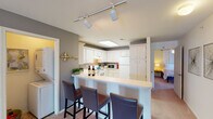 This single-floor unit at Varsity features three bedrooms and three full bathrooms within a well-organized layout. The living room is finished with neutral gray walls, plush carpet flooring, and windows that allow natural light to filter in, complemented by a direct exterior access door. Seating areas flow naturally into the adjoining kitchen, which is separated by a breakfast bar with counter seating for three—ideal for casual dining. The kitchen itself is equipped with white cabinetry offering ample storage, a full suite of white appliances including an electric range and refrigerator, a skylight panel for added brightness, and track lighting overhead.

Each of the three bedrooms features carpeted floors, ceiling fans with light fixtures, and windows providing natural light and outdoor views. The bedrooms vary slightly in size but each offers a comfortable, functional space. All three bathrooms include a single-sink vanity with under-sink storage, a large wall-mounted mirror with overhead lighting, and a shower-tub combination. Tile flooring is consistent throughout the bathrooms. In-unit laundry connections are also visible, adding everyday convenience to this well-appointed multi-bedroom layout.
