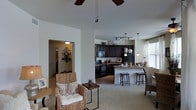 This single-floor home showcases a thoughtfully designed open-concept floor plan that effortlessly connects the main living areas. The living room is warm and inviting, featuring a ceiling fan overhead and neutral-toned walls that create a bright, airy atmosphere. Natural woven accent chairs add a relaxed, coastal-inspired touch to the seating area.

The kitchen is a standout feature, boasting rich dark cabinetry that contrasts beautifully with lighter countertops. A central island with a raised breakfast bar provides additional seating and serves as a natural gathering point. The kitchen is equipped with black appliances, a gas range, and overhead track lighting, while pendant lights illuminate the island below. Ample upper and lower cabinet storage ensures functional organization throughout.

Adjacent to the kitchen, a dedicated dining area benefits from natural light streaming in through a nearby window, enhancing the home's overall warmth. A hallway visible from the living room leads to additional spaces within the home, hinting at a well-planned layout designed for comfortable everyday living. Decorative wall accents and carefully selected furnishings throughout contribute to a cohesive, welcoming aesthetic that balances style with practicality.
