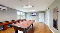 This single-floor game room is a well-appointed recreational space designed for leisure and social activity. The room is finished with warm hardwood flooring throughout, complementing the neutral gray-toned walls that give the space a clean, contemporary feel. At the heart of the room sits a full-size pool table with a classic felt surface, accompanied by a wall-mounted cue rack keeping equipment neatly organized and accessible. A shuffleboard table runs along one side of the room, adding an additional layer of entertainment options for guests. A wall-mounted flat-screen television provides an added amenity, making the space suitable for watching games or casual viewing while playing. Overhead, recessed panel lighting ensures the area is well-lit throughout. Large sliding windows along one wall flood the room with natural light and offer views of lush, tree-lined grounds outside. A split-system air conditioning unit provides climate control for year-round comfort. Glass-paned double doors connect the game room to adjacent common areas, enhancing its open and sociable character. Restroom access is conveniently located within the space. Overall, this game room delivers a functional and inviting environment ideal for community recreation and entertainment.