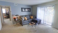 This single-floor space offers a comfortable and modern living environment designed with both style and functionality in mind. The living room serves as the focal point of the space, showcasing a soothing blue-gray color palette that creates a calm and inviting atmosphere. Plush wall-to-wall carpeting adds warmth underfoot, while large sliding glass doors fitted with vertical blinds flood the room with abundant natural light and provide access to the outdoors. The room is thoughtfully arranged with a contemporary sectional sofa in charcoal gray, accented with decorative throw pillows in warm mustard and patterned tones, complementing the neutral color scheme. A sleek glass-top coffee table and a matching side table add an elegant, airy feel to the space without overwhelming it visually. A framed black-and-white cityscape artwork anchors the main wall, adding a sophisticated urban touch. Overhead ventilation is integrated seamlessly into the ceiling, reflecting a practical and well-maintained interior. The overall ambiance is clean, bright, and modern, making it an inviting space suited for both relaxing and entertaining.