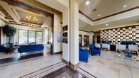This well-appointed single-floor facility showcases a refined Mediterranean-inspired design, blending warm neutral tones, rich dark wood accents, and stone tile flooring throughout. The grand entrance features floor-to-ceiling glass double doors with transom windows, flooding the foyer with natural light and offering views of the surrounding landscaping.

The main lobby is anchored by soaring tray and coffered ceilings detailed with crown molding, complemented by elegant chandelier lighting and recessed fixtures. Structural columns clad in dark wood framing define the transition between spaces while maintaining an open, airy feel. Bold cobalt blue upholstered seating is arranged thoughtfully across multiple lounge areas, creating inviting spaces for gathering and relaxation.

A sunlit sitting room boasts an expansive bank of gridded windows overlooking lush outdoor greenery, with a decorative chandelier and large potted trees enhancing the organic ambiance. A central lounge area features a wall-mounted TV above a sleek linear fireplace set against a tiled accent wall. The facility also includes a fully equipped kitchen with dark cabinetry, stainless steel appliances, a white quartz island with bar seating, and a built-in TV. Additional quieter seating nooks with carpeted flooring and curated wall art provide more intimate settings throughout the space.