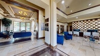 This well-appointed single-floor facility showcases a refined Mediterranean-inspired design, blending warm neutral tones, rich dark wood accents, and stone tile flooring throughout. The grand entrance features floor-to-ceiling glass double doors with transom windows, flooding the foyer with natural light and offering views of the surrounding landscaping.

The main lobby is anchored by soaring tray and coffered ceilings detailed with crown molding, complemented by elegant chandelier lighting and recessed fixtures. Structural columns clad in dark wood framing define the transition between spaces while maintaining an open, airy feel. Bold cobalt blue upholstered seating is arranged thoughtfully across multiple lounge areas, creating inviting spaces for gathering and relaxation.

A sunlit sitting room boasts an expansive bank of gridded windows overlooking lush outdoor greenery, with a decorative chandelier and large potted trees enhancing the organic ambiance. A central lounge area features a wall-mounted TV above a sleek linear fireplace set against a tiled accent wall. The facility also includes a fully equipped kitchen with dark cabinetry, stainless steel appliances, a white quartz island with bar seating, and a built-in TV. Additional quieter seating nooks with carpeted flooring and curated wall art provide more intimate settings throughout the space.
