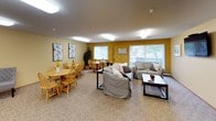 This single-floor space offers a warm and inviting atmosphere, characterized by a cohesive color palette of soft cream and bold golden-yellow accent walls that create a cheerful, communal ambiance throughout. The main area is a generously sized open room that seamlessly combines a living lounge and a dining area. The lounge side is furnished with comfortable upholstered sofas arranged around a central coffee table, with a large flat-screen television mounted on the wall for easy viewing. Multiple windows fitted with vertical blinds allow natural light to filter into the space, offering views of the greenery outside. The dining area accommodates multiple round wooden tables with matching Windsor-style chairs, creating a casual and sociable setting ideal for shared meals or gatherings. A decorative indoor plant and framed artwork on the walls add a homey, curated touch to the dining space. The kitchen is compact yet well-equipped, featuring warm oak cabinetry, white laminate countertops, a double-basin sink, a coil electric range, and ample upper and lower cabinet storage. Wood-look flooring in the kitchen contrasts with the textured carpet found throughout the main living and dining areas. Flush-mount ceiling lighting provides consistent illumination across all areas, complementing the natural light from the windows.