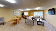 This single-floor space offers a warm and inviting atmosphere, characterized by a cohesive color palette of soft cream and bold golden-yellow accent walls that create a cheerful, communal ambiance throughout. The main area is a generously sized open room that seamlessly combines a living lounge and a dining area. The lounge side is furnished with comfortable upholstered sofas arranged around a central coffee table, with a large flat-screen television mounted on the wall for easy viewing. Multiple windows fitted with vertical blinds allow natural light to filter into the space, offering views of the greenery outside. The dining area accommodates multiple round wooden tables with matching Windsor-style chairs, creating a casual and sociable setting ideal for shared meals or gatherings. A decorative indoor plant and framed artwork on the walls add a homey, curated touch to the dining space. The kitchen is compact yet well-equipped, featuring warm oak cabinetry, white laminate countertops, a double-basin sink, a coil electric range, and ample upper and lower cabinet storage. Wood-look flooring in the kitchen contrasts with the textured carpet found throughout the main living and dining areas. Flush-mount ceiling lighting provides consistent illumination across all areas, complementing the natural light from the windows.