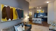 This single-floor space offers a thoughtfully designed open-concept layout that seamlessly connects the living, dining, and kitchen areas into one cohesive living environment. Soaring ceilings lend an impressive sense of height and volume, enhancing the overall spaciousness of the interior.

The living area is anchored by a plush gray sofa set atop a soft area rug, complemented by a large, bold abstract artwork that adds a vibrant pop of color against the cool gray walls. Track lighting and a stylish table lamp provide warm, layered illumination throughout.

The kitchen is a standout feature, boasting dark cabinetry, stainless steel appliances, and a sleek white kitchen island with bar seating — ideal for casual dining or entertaining. A tile backsplash and modern countertops complete the polished look. Adjacent to the kitchen, a cozy dining nook with a pendant light fixture offers an intimate space for meals.

Rich hardwood-style flooring flows throughout the space, tying together the modern aesthetic with warmth and continuity. The cohesive color palette of charcoal gray, white, and yellow accents — echoed in both the artwork and decor — gives the space a curated, contemporary character that is both inviting and visually dynamic.