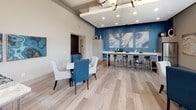 Located at 350 Bradley Park Lane in Cumming, Georgia, this single-story community clubhouse offers a thoughtfully designed, open-concept gathering space that balances comfort with contemporary style. Light hardwood flooring runs throughout, providing a cohesive foundation that ties together each functional zone.

The lounge area is a standout feature, anchored by a generously sized sectional sofa in neutral tones accented with teal and blue throw pillows. Soaring ceilings with transom windows along two walls flood the space with abundant natural light and frame views of the surrounding community. A wall-mounted flat-screen television is paired with a decorative mirrored media console, while curated artwork in blue and gold tones adds visual interest.

The social and work zone features a bold blue textured accent wall serving as a dramatic backdrop for a series of large abstract art panels. A long white bar-height counter with seating accommodates both collaborative work and casual socializing. Dining tables with a mix of white and blue upholstered chairs provide additional flexible seating.

The kitchenette is well-appointed with white cabinetry, a stainless steel sink, refrigerator, and a professional-grade espresso and coffee station, making this space ideal for resident gatherings and events. The overall ambiance is modern, airy, and welcoming.