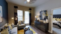 This single-floor residence showcases a thoughtfully designed living area that blends contemporary style with comfortable functionality. The living room features rich hardwood-style flooring that anchors the space, complemented by neutral warm-toned walls that create an inviting atmosphere. A ceiling fan overhead provides both practical airflow and a subtle design element.

Natural light filters generously into the room through a large window and a glass-paneled door, dressed with bold dark curtains featuring decorative patterned valances that add a dramatic contrast to the lighter walls. The window treatments frame views of the surrounding urban exterior, hinting at a vibrant neighborhood setting.

The space is furnished with a cohesive, modern aesthetic — a light gray sofa and accent chair are paired with round coffee tables and a yellow geometric-patterned area rug that adds a pop of color. A dark-toned dresser serves as a media and display console, topped with curated décor including a table lamp, vases, and a large abstract artwork in blue and gold tones. A multi-arm floor lamp provides additional ambient lighting, completing the polished, urban-contemporary feel of the space.