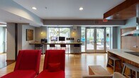 This single-floor amenity space is thoughtfully designed to offer residents a variety of social, recreational, and functional zones within one expansive layout. The lounge area serves as a natural centerpiece, anchored by a striking floor-to-ceiling stacked-stone fireplace with a built-in linear hearth and a large wall-mounted television above it. Bold red seating adds a vibrant pop of color against the warm hardwood floors and neutral gray walls throughout.

The fully equipped kitchen features rich bamboo cabinetry, a dramatic wood-paneled ceiling, stainless steel appliances, a large island with a stone composite countertop, a cooktop with an overhead range hood, and bar stool seating — ideal for entertaining or communal cooking. Adjacent dining areas include high-top red tables with contemporary bar chairs set against teal accent walls and large-scale abstract artwork.

A dedicated business center provides computer workstations and a printer beneath pendant lighting, with floor-to-ceiling glass doors that open directly to an outdoor patio, filling the space with natural light and greenery views. The billiards area rounds out the recreational offerings. Together, these spaces create a dynamic, well-lit environment that balances productivity, relaxation, and community engagement.
